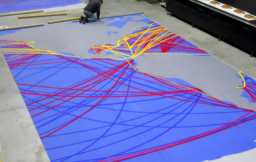 A large printed carpet of a world map with route arcs, laid out flat while a worker checks the seam transitions between panels
